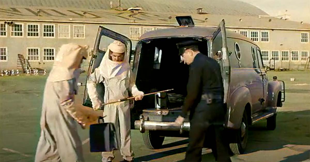 Two scientists in protective suits and a uniformed officer load a cylindrical container into the back of a dark-colored transport vehicle.