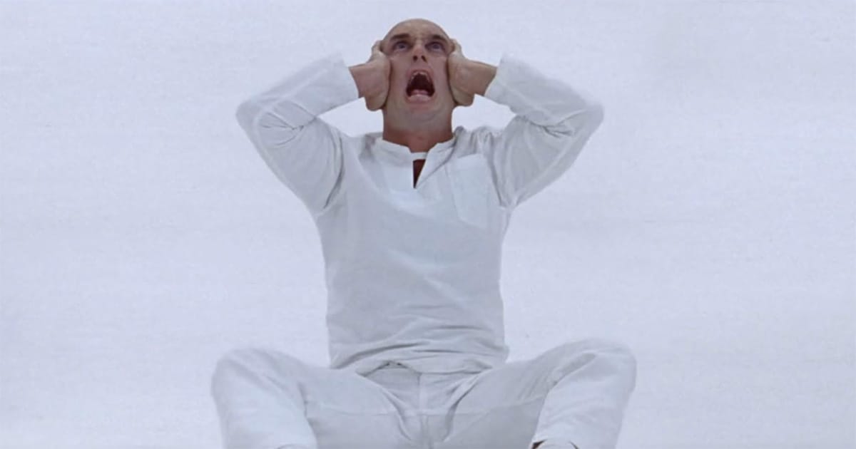 A bald man in a white outfit sits on the floor, clutching his head and screaming in a stark white void.