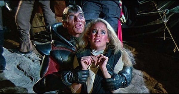 Jared-Syn, in black armor, holds Shala, a frightened woman, in dimly lit scene from Metalstorm: The Destruction of Jared-Syn.