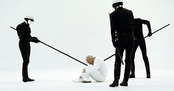 A bald man in white sits as three black-clad enforcers stand over him with batons.